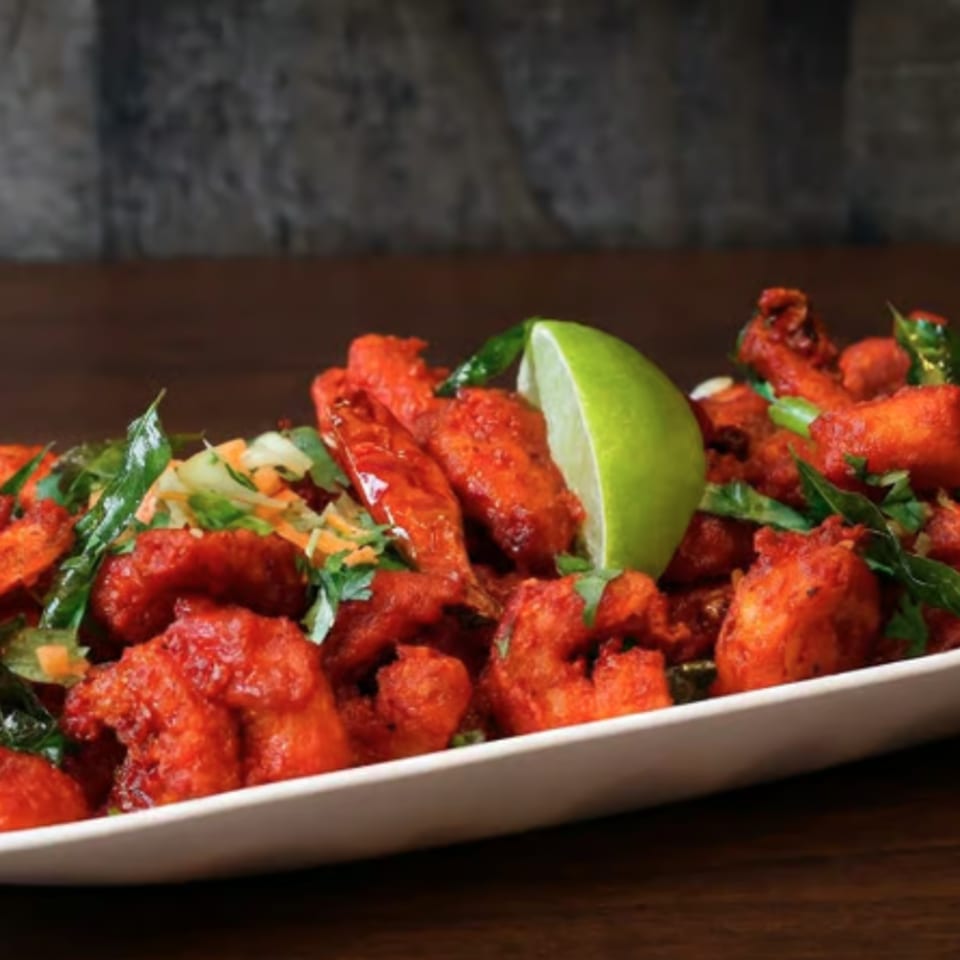 Plate of crispy red fried chicken pieces garnished with curry leaves, herbs, and a lime wedge on a wooden table.