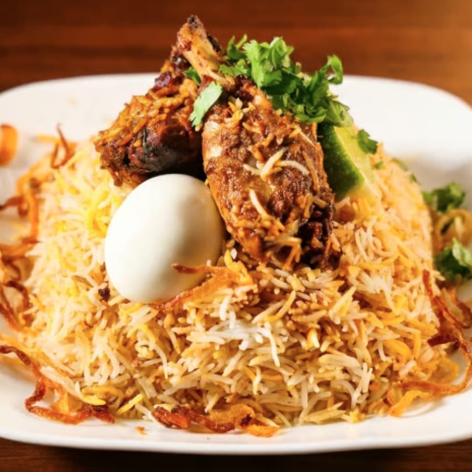 Plate of fragrant biryani rice topped with fried chicken, boiled egg, cilantro, cucumber, and crispy onions.