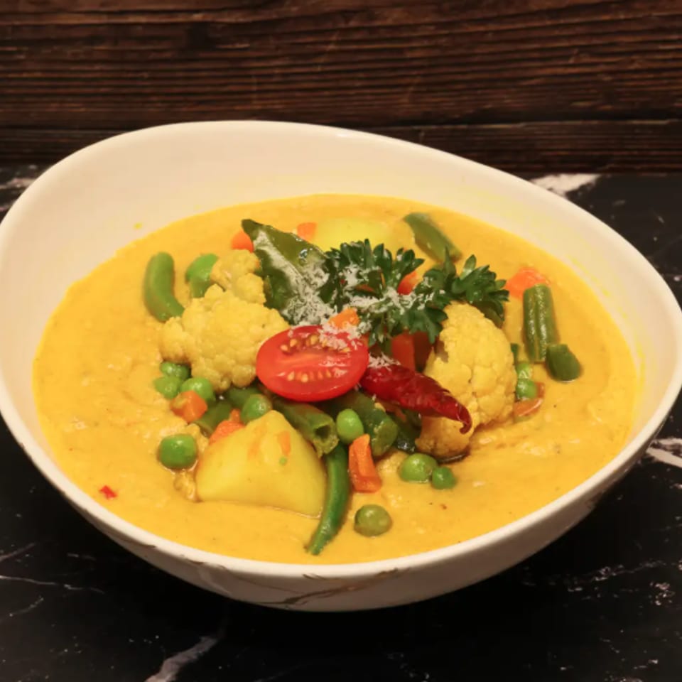 Bowl of creamy yellow vegetable curry topped with cauliflower, peas, peppers, herbs, tomato, and red chili.