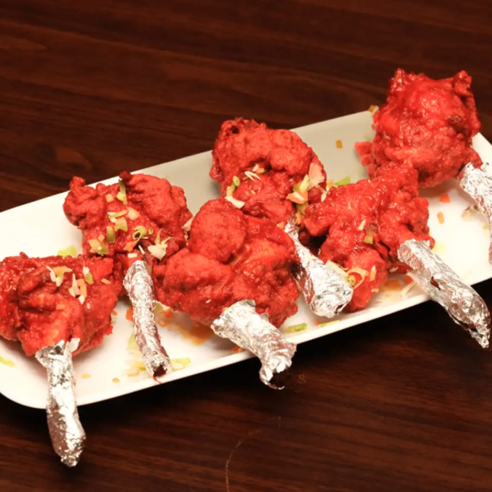 Plate of bright red, garnished chicken lollipops with foil-wrapped handles on a dark wooden table.