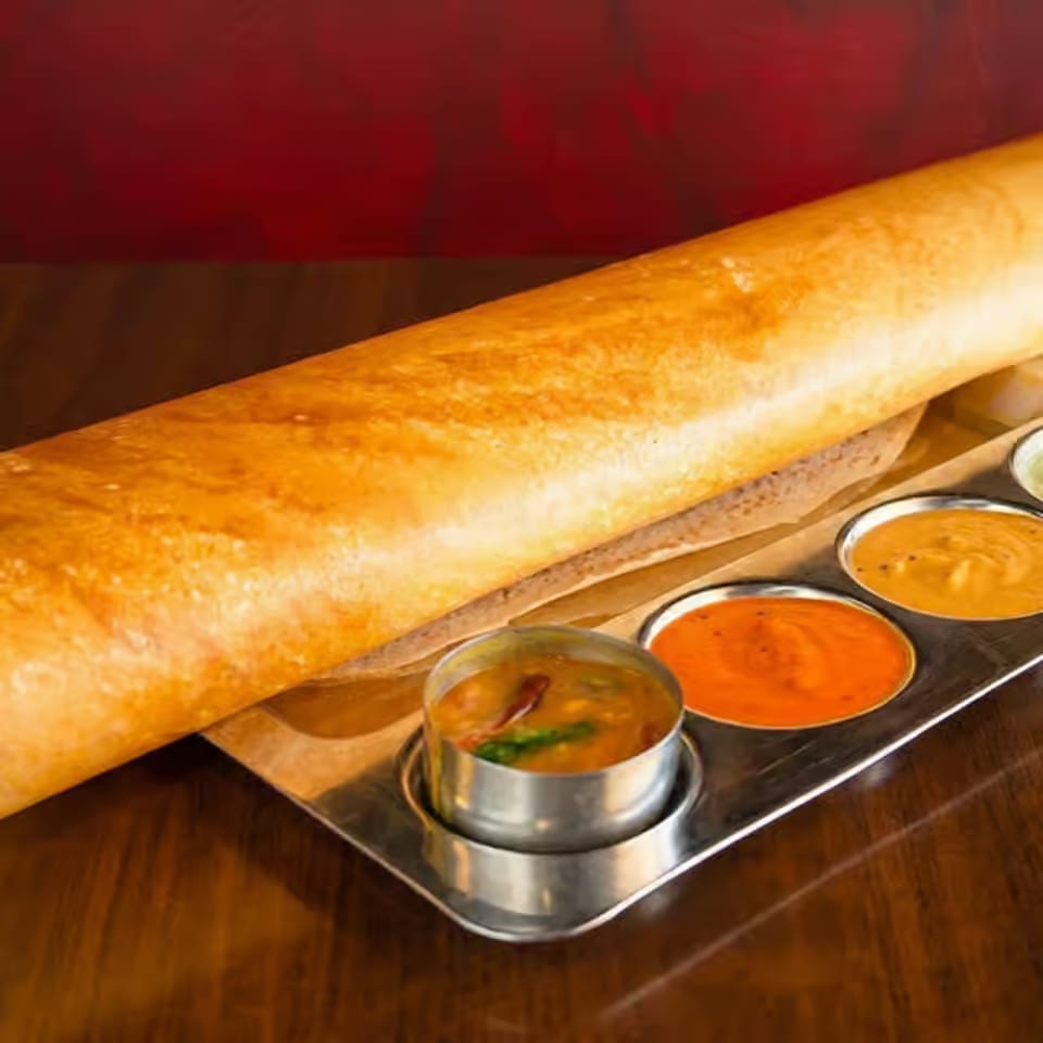 Crispy golden dosa served on a wooden board with three small bowls of colorful chutneys and sambar.