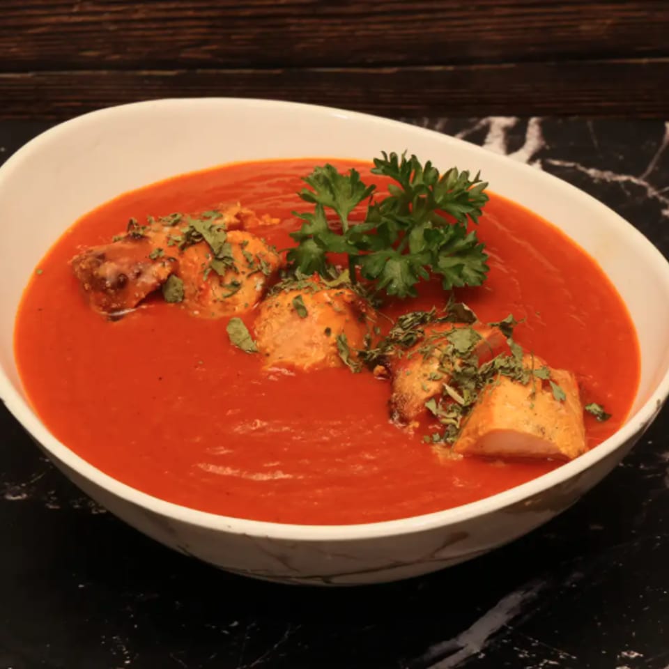 Bowl of vibrant red tomato soup topped with chicken pieces, chopped herbs, and fresh parsley on a black marble table.