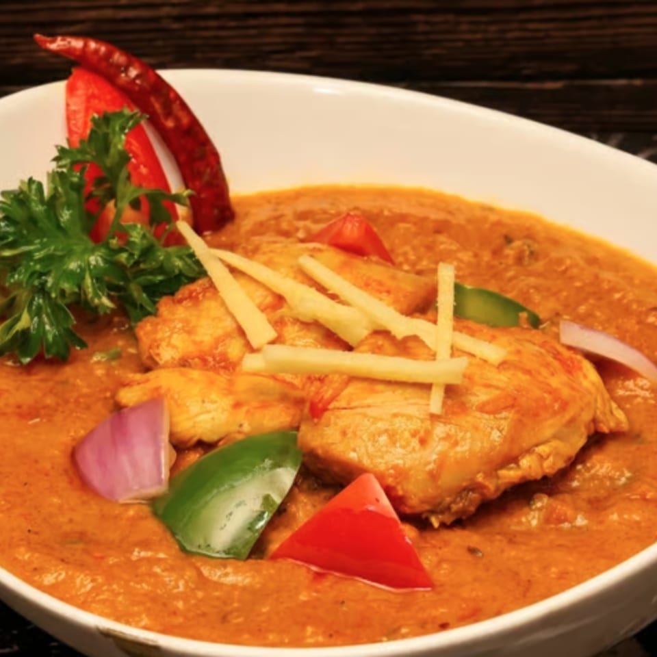 Bowl of creamy orange curry with grilled chicken, sliced vegetables, herbs, and crispy garnish on a dark table.